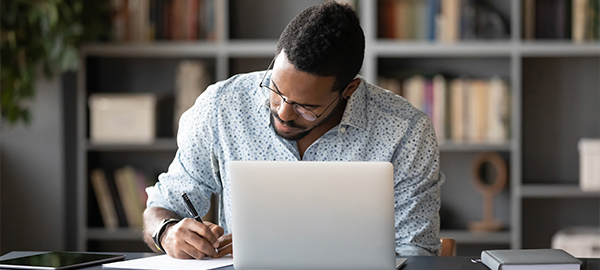 A man using a pen to write while sitting in front of a laptop computer.