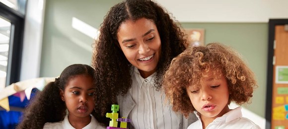 A teacher is between two children as the three engage in a learning activity that's just out of frame of the photo. The teacher smiles as the children share an inquisitive look.