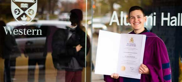 An alum holds up their degree in front of Alumni Hall at Western University.