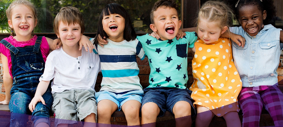 Six children sit on a bench while smiling and posing for the camera.