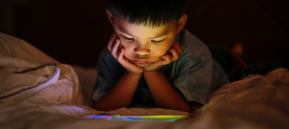 A child lays in bed with his eyes fixated on a mobile electronic device.