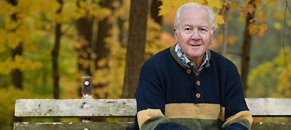 Photo of John Davidson sitting on a park bench in a warm sweater in the fall